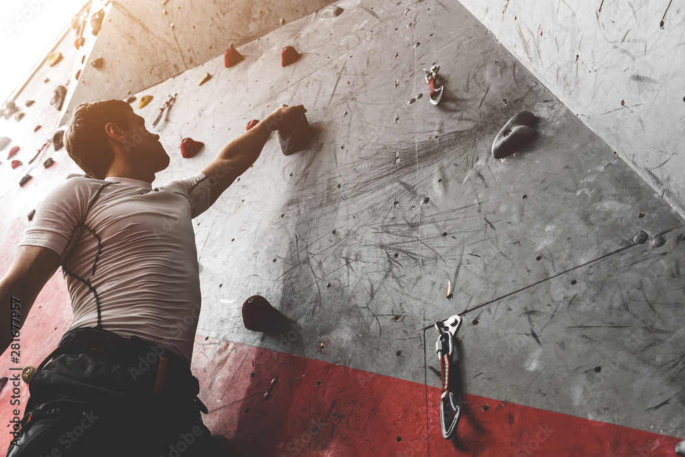 Wall mural Sportsman climber moving up on steep rock, climbing on artificial wall indoors. Extreme sports and bouldering concept