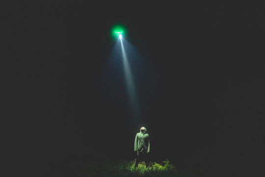 The UFO shines on a male standing on the grass. night time