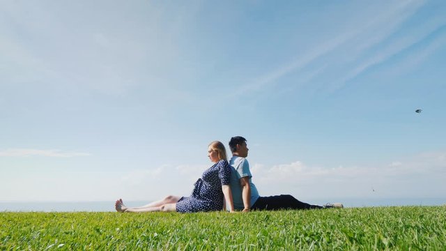Asian man with a pregnant woman are resting in nature in a picturesque place, sit on the green grass