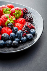 A vertical shot of a plate of fresh summer fruit showing strawberries blue berries black berries and mulberries on a dark background.