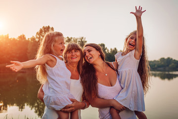 Family walking by summer river at sunset. Mother and grandmother holding kids and laughing. Three denerations