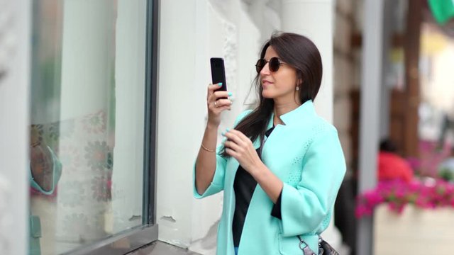 Smiling beautiful woman admiring taking photo of glass shop showcase using smartphone
