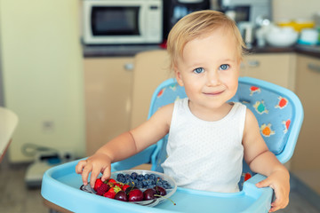 Adorable cute caucasian blond toddler boy enjoy tasting different seasonal fresh ripe organic berries sitting in highchair at home kitchen. Happy kid eating natural sweet healthy food as lunch snack