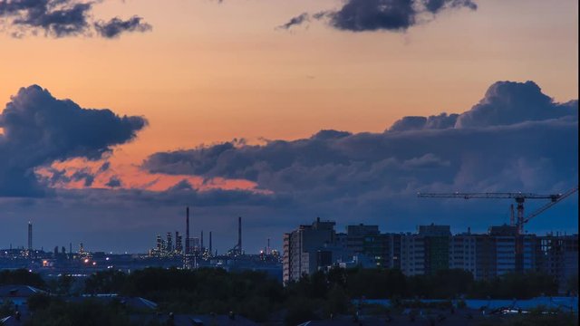 Epic storm clouds moving fast at sunset over city skyline 4K time lapse. Oil refinery lights, power plant pipes with smoke. Constraction site with big crane