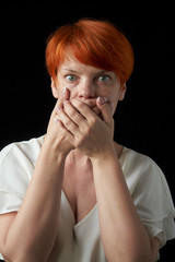Adult red-haired woman closed her mouth with her hand because she was surprised or scared, rubbed on a black background, selective focus