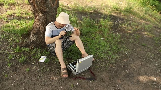 young caucasian scientist entomologist with glasses and panama hat sits under a tree and examines beetles in a bank, medium plan