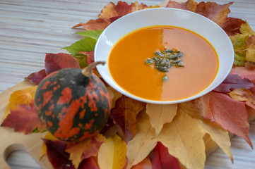 Tasty pumpkin bright orange soup with green seeds served on white plate and autumnal colorful leaves