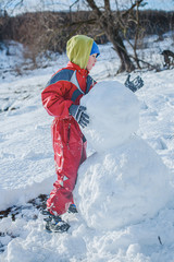 boy in red jumpsuit builds snowman out of snow against background of forest. Heavy snowfall with sleet. child plays active games outside in winter.