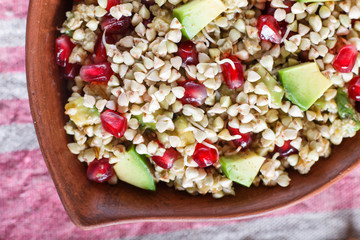 Salad of germinated buckwheat, avocado, walnut and pomegranate seeds in clay plate on white wooden background.