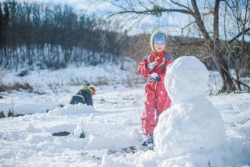 boy in red jumpsuit builds snowman out of snow against background of forest. Heavy snowfall with sleet. child plays active games outside in winter.