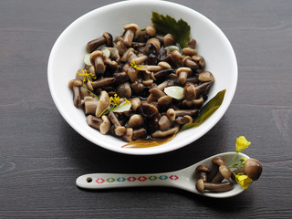 Delicious, beautiful, small pickled mushrooms in a dessert spoon close-up. Home recipe - delicious pickles with garlic, pepper, Bay leaf, cherry leaf and herbs in a white plate on a wooden table.