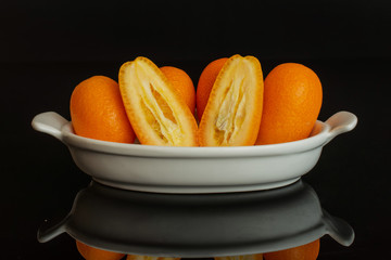 Group of four whole two halves of fresh orange kumquat in a white oval ceramic bowl isolated on black glass