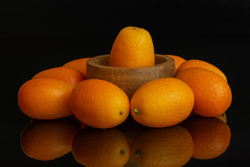 Lot of whole fresh orange kumquat placed around the one that is in a tiny wooden bowl isolated on black glass