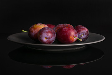 Group of five whole fresh blue plum on a gray ceramic plate isolated on black glass