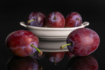 Group of five whole fresh blue plum in a white oval ceramic bowl isolated on black glass