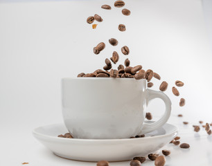 Coffee beans fall into a white porcelain cup and saucer, bouncing and jumping through the air conveying alert excitement, all isolated against a white background