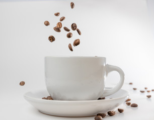 Coffee beans fall into a white porcelain cup and saucer, bouncing and jumping through the air conveying alert excitement, all isolated against a white background