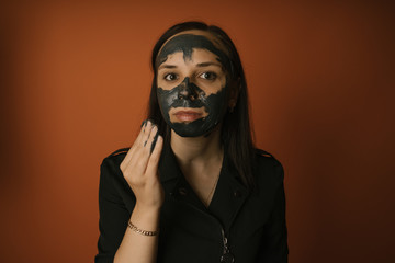 A young woman inflicts a black mask on the face on an orange background. The concept of healthy lifestyle, beauty, body care.