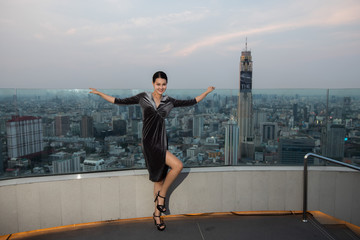 Asian woman in evening dress stand on top roof of Hotel in cityscape city view with horizontal twilight period
