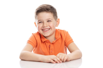 Laughing school-age boy in a bright orange polo t-shirt sits at a table. Close-up. Isolirvoan on a white background.