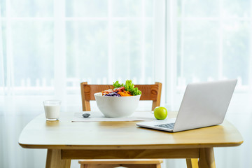 Scene of vegetables in the white bolw with a milk of glasses and apple with technology laptop on the wooden table with chair in the moderm home, Healthy and work hard concept