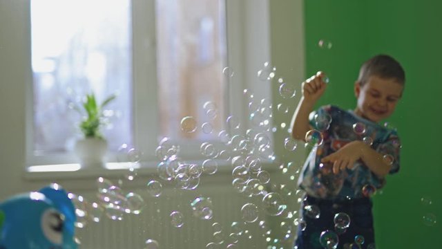 Joyful boy plays with soap bubbles in his room. Little child is popping colorful bubbles with his fingers and smiling at home.