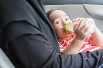 One year old Thai baby boy drink milk from a bottle in a car with mother hugging.
