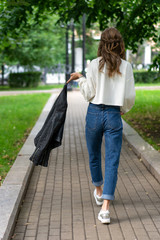 Beautiful young European girl posing on sidewalk in city green park. Caucasian girl skip back. Lifestyle, pullover, jeans, sneakers. interesting pose with your back full length in trendy look.