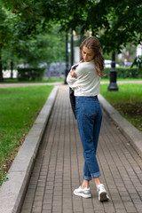 Fototapeta premium Beautiful young European girl posing on sidewalk in city green park. Caucasian girl skip back. Lifestyle, pullover, jeans, sneakers. interesting pose with your back full length in trendy look.