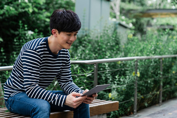 Relaxed Asian man in casual clothes using tablet while sitting on a bench in a park.