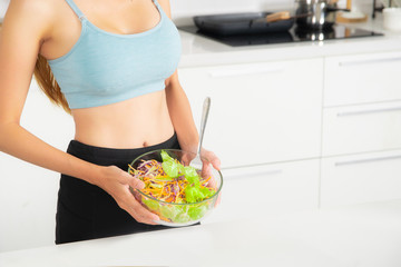 Shapely girl and love health wearing a blue fitness suit, black pant standing holding glass bowl with salad at modern white kitchen, close-up view.