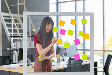 Beautiful asian woman writing colorful paper on clear glass memo board in office