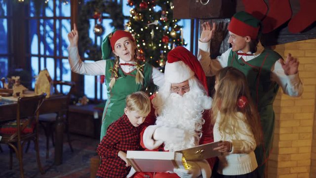 Santa with little kids and elves dancing on Christmas background. Santa Claus is showing photo album to children while joyful elves having fun inside the house.