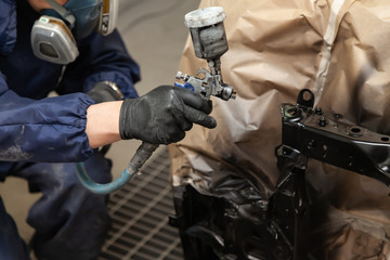 A male worker in jumpsuit and gloves paints with a spray gun a front frame part of the car body in black after being damaged at an accident. Auto service industry professions