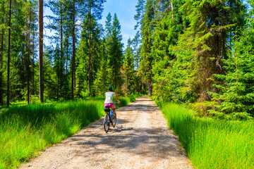 Young woman riding bike in forest on cycling way around Tatra Mountains on sunny summer day, Poland