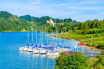 Sailing boats in port on shore of Czorsztynskie lake with castle in background on beautiful summer day in Niedzica village, Pieniny Mountains, Poland