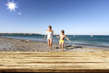 Wooden table background with seaside and family view. Mother and her son playing and having fun on the beach.