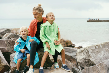 Fototapeta premium Two sons in raincoats with mom in rainy weather on the seashore. Joint pastime, family values, close relationships.