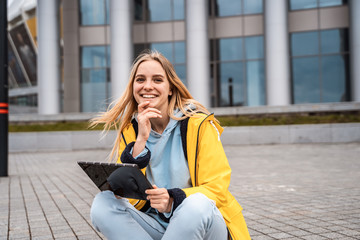 A beautiful blonde girl sits on a street and uses a tablet.