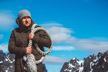 Fototapeta na wymiar Brutal man with a rope on his shoulder against the background of the mountains and the blue sky. Copy space. Can use as banner