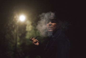 Detective agent in a hat and coat smokes his smoking pipe and thinking in a night park.