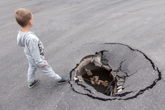 A child stands on the brink of a large pit in the asphalt. Danger of injury to children on a city street. Road surface hole