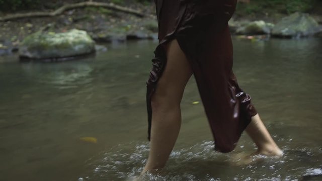 Beautiful pregnant woman in a dark brown dress goes barefoot on water in the tropical jungle river. Slow motion.