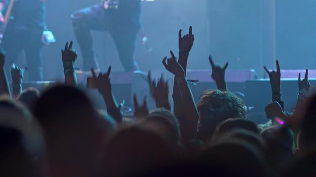 People in a crowd jumping to the groove during a rock concert. They have their hands showing horns gesture. The stage is illuminated with blue light while a band is performing. 4K