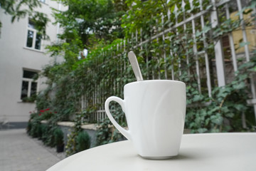 white Cup with coffee, tea or cocoa on a saucer with a spoon on the summer terrace, against the urban landscape. the fence is entwined with greenery and tall buildings.
