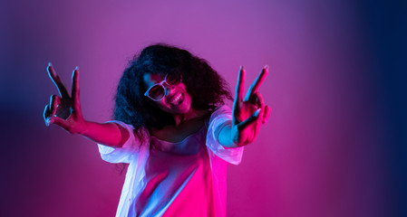 Fashion young african girl black woman wear stylish pink glasses dance look at camera show peace gesture enjoying festive party isolated on disco purple studio background, portrait, copy space