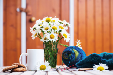 Women's hobby. Crochet and knitting. Workspace. A bouquet of daisies in a vase, a white Cup with a drink, cookies, yarn balls, needles, scissors, knitwear, crochet hooks on a wooden table in a home