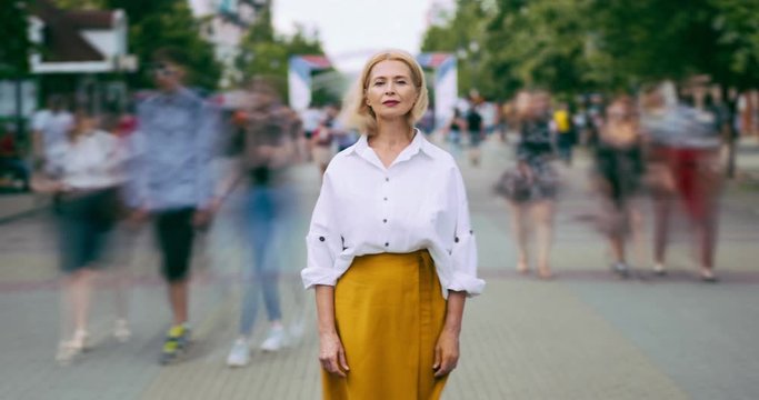 Time lapse portrait of beautiful mature lady with serious face in city street standing alone looking at camera. Summer town, people and modern life concept.