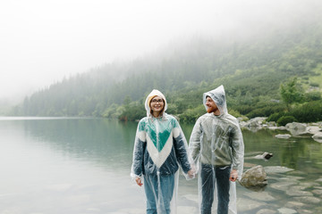 Fototapeta premium Outdoor shot of young couple in love wearing rain coats and standing in front of beatiful lake in mountains. Beautiful couple posing on nature landscape