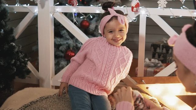 Happy mother having fun with her kid daughter near Christmas tree. Mother tickling her little daughter. Beautiful mother and daughter in pink pullovers at Christmas day. Happy family concept